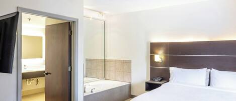 Una habitació d'hotel amb un llit, una taula de nit, un mirall, un bany amb banyera i una porta que condueix a una altra habitació.