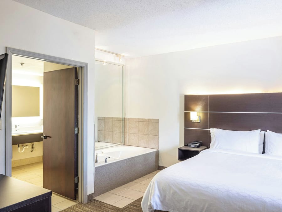 Habitación de hotel con cama, mesita de noche, espejo, baño con bañera y puerta que da a otra habitación.