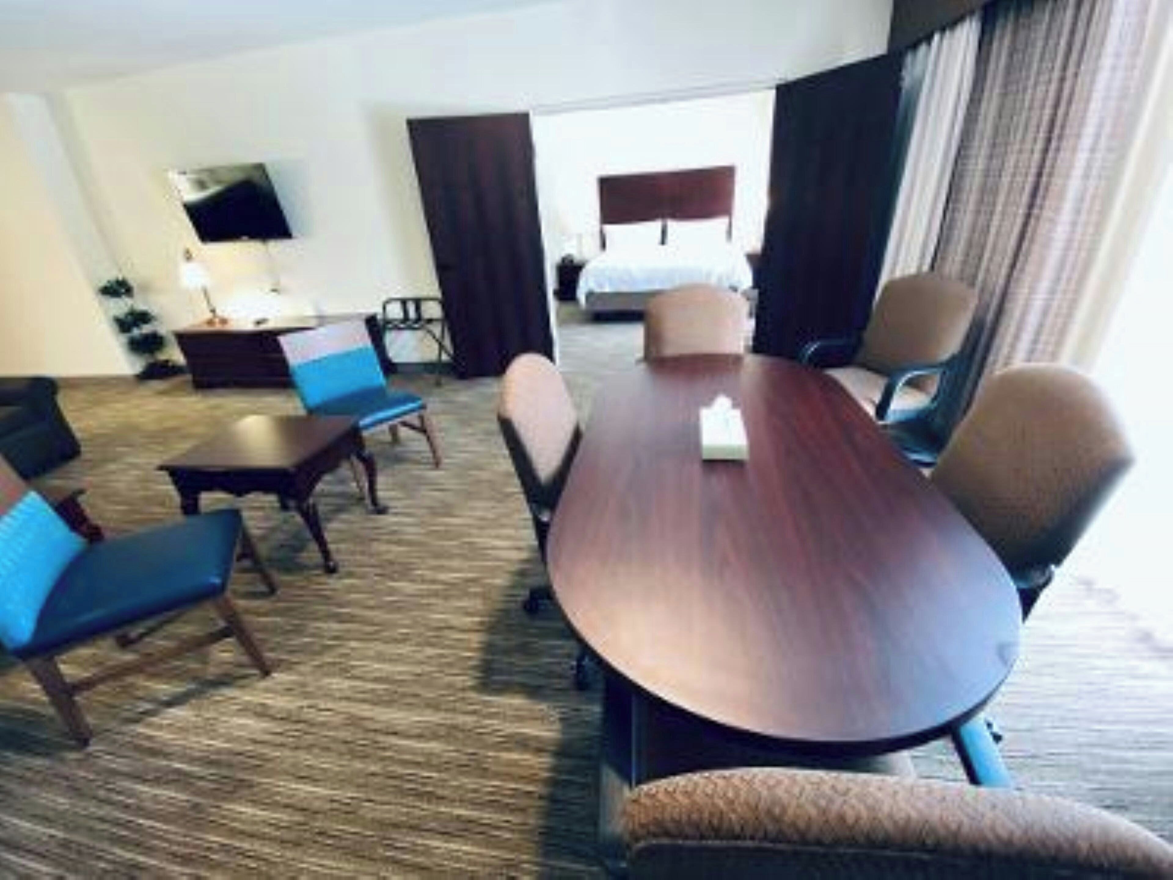 A hotel room with a large wooden table, several chairs, a sofa, a TV mounted on the wall, and a bed with a headboard.