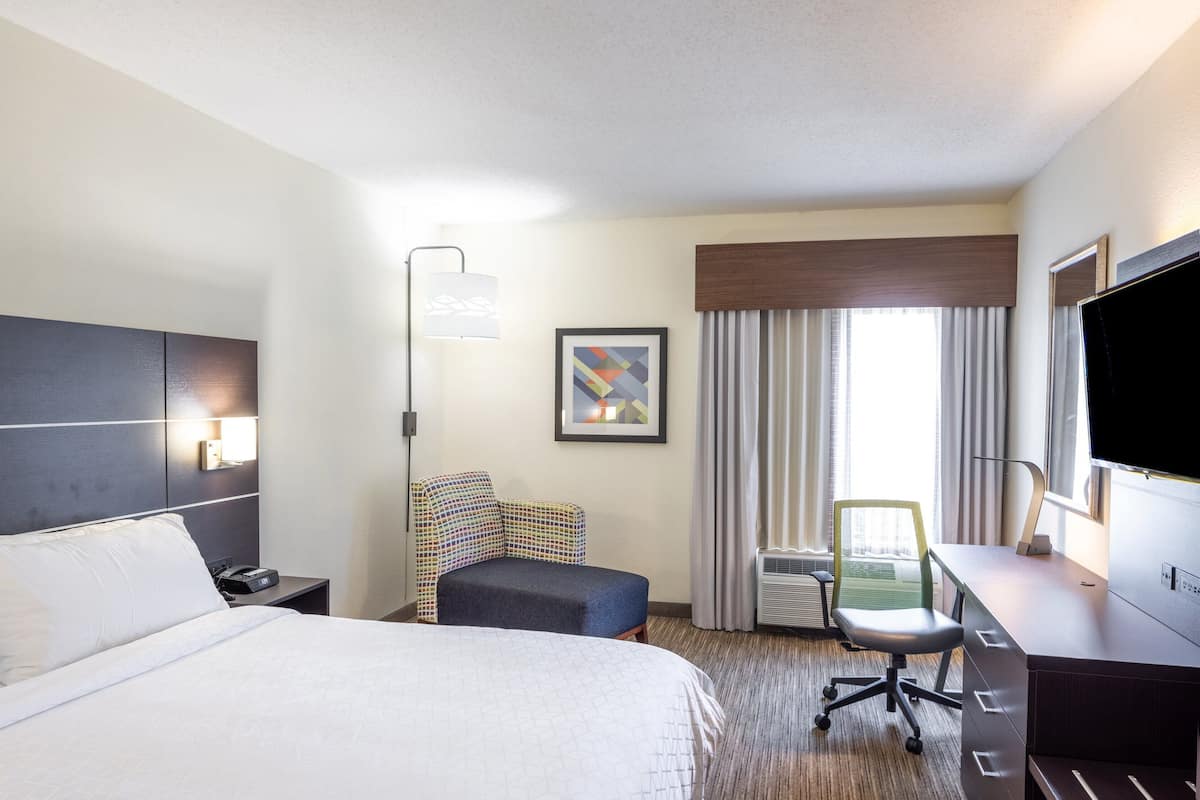 A hotel room with a large bed, a desk with a chair, a flat-screen TV, a window with curtains, and a framed picture on the wall.