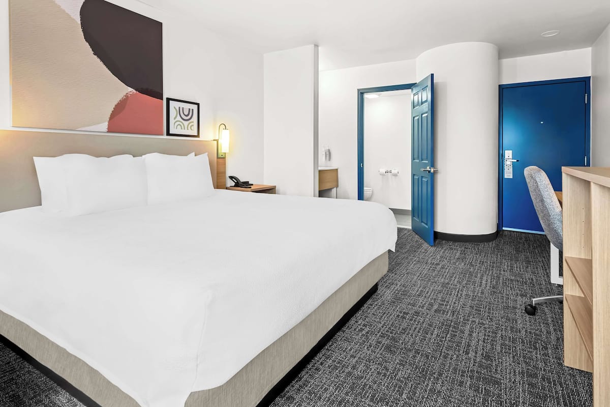 A hotel room with a large bed, a desk, a chair, a bathroom, and a blue door.