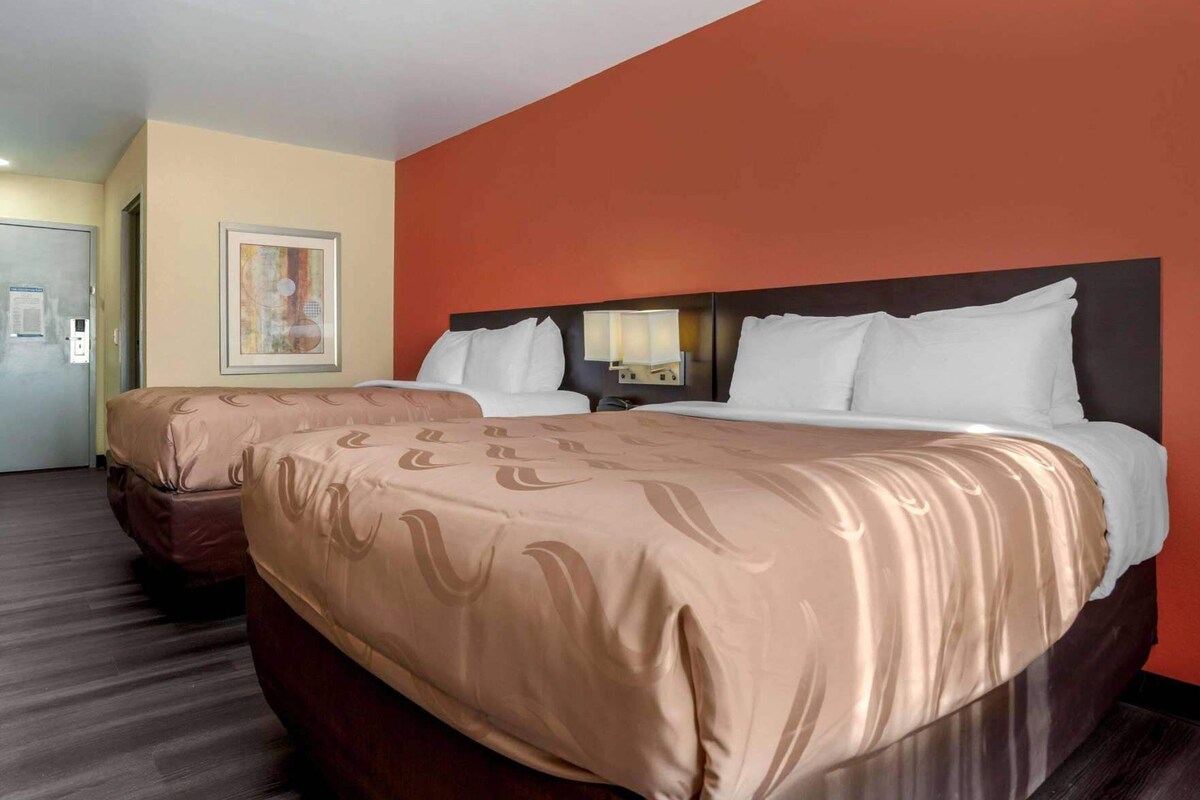 Une chambre d’hôtel avec deux lits, un mur d’accent rouge, une œuvre d’art encadrée et une porte sur le côté gauche.