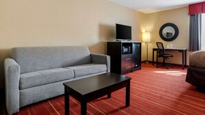Habitación de hotel con sofá gris, una mesita, televisor en un mueble bajo, escritorio con silla y una cama.