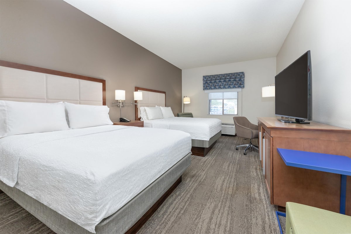 A hotel room with two beds, a desk, a television, and a window.