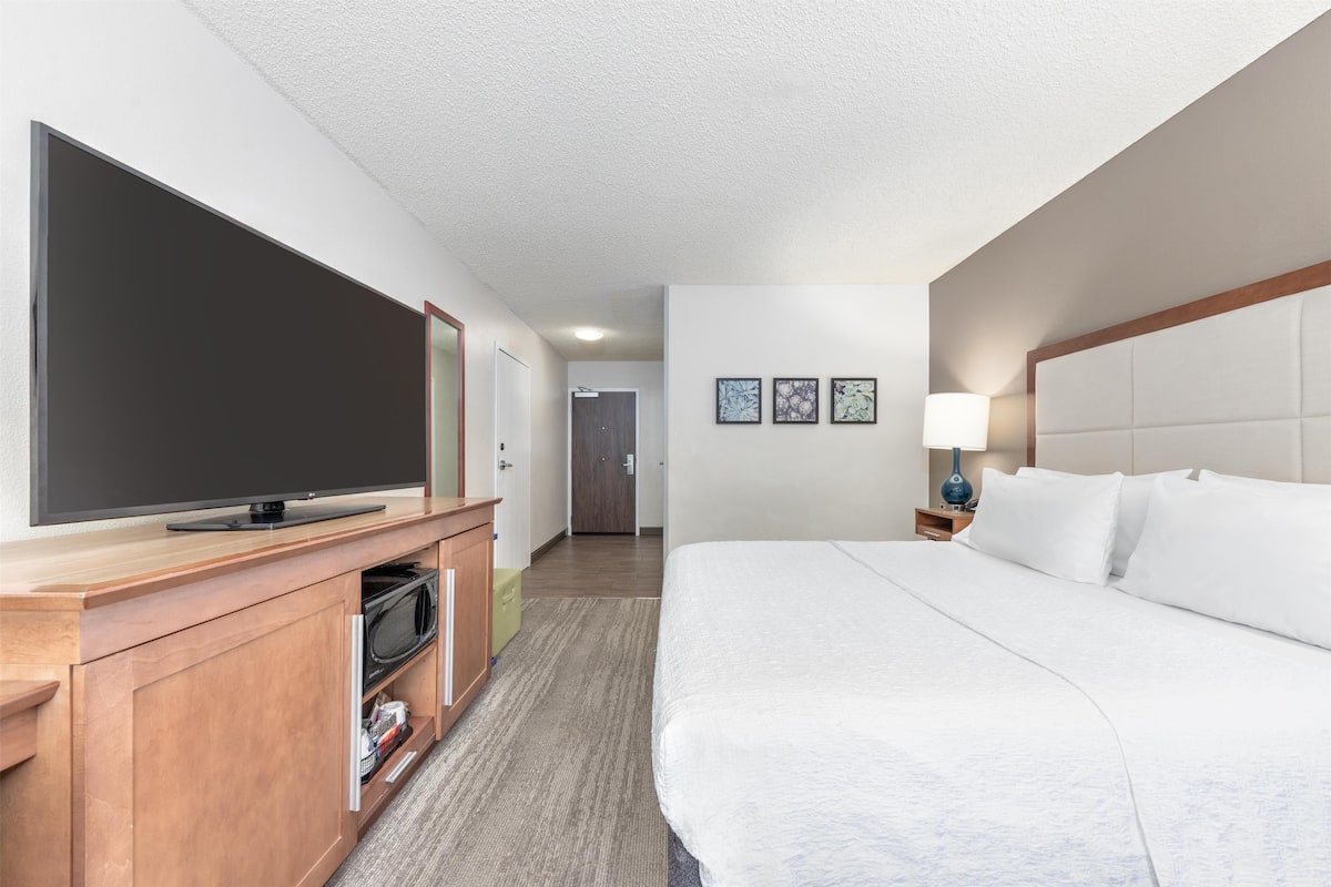 A hotel room with a large flat-screen TV, a bed with white linens, a wooden dresser, and a nightstand with a lamp.