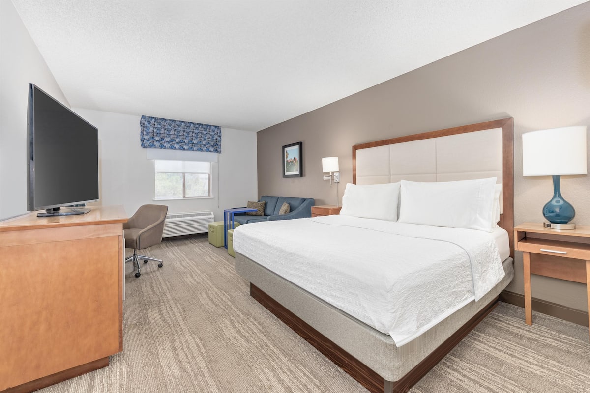 A hotel room with a bed, desk, chair, TV, and a window with blue curtains.