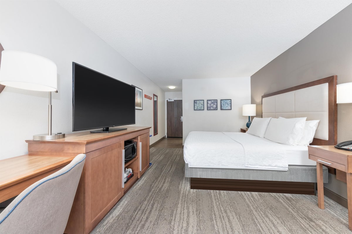 A hotel room with a bed, a desk with a television, a chair, and a bedside table.