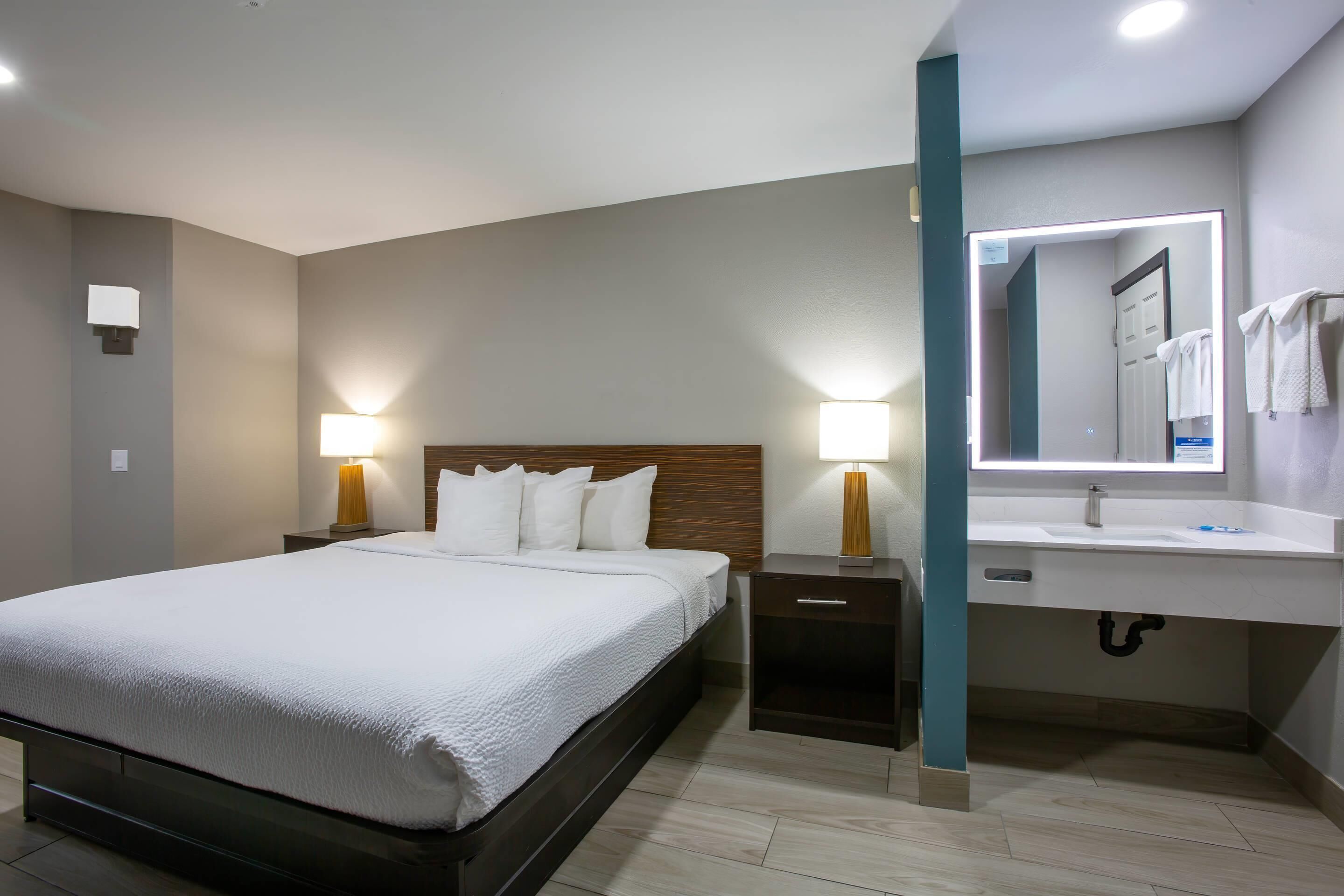 Quarto de hotel com cama, abajures, mesa de cabeceira, painel de cabeceira de madeira, banheiro com pia e espelho, e toalhas em um suporte.