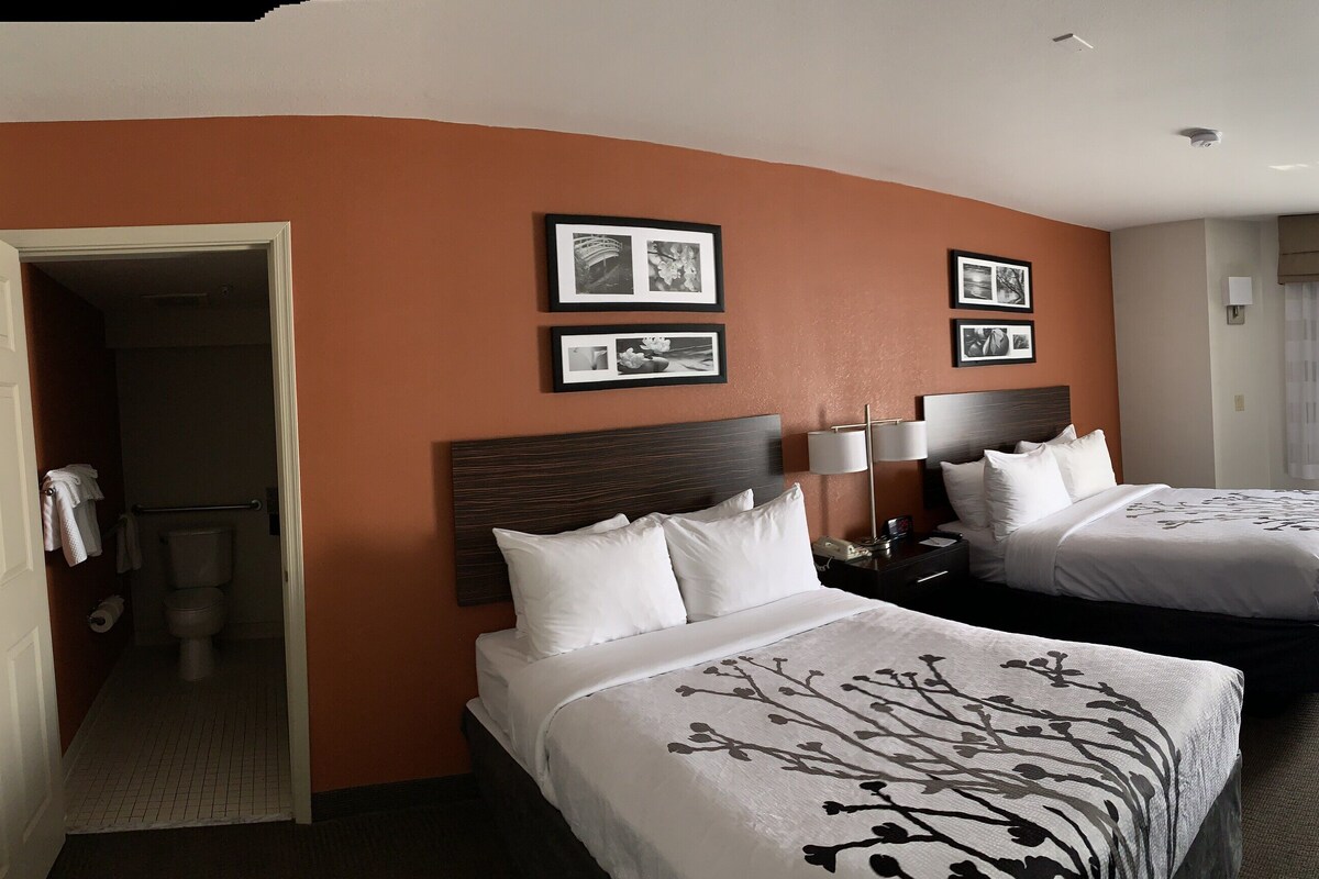 Ein Hotelzimmer mit zwei Betten, einem Fernseher, einem Schreibtisch und einem durch eine offene Tür einsehbaren Badezimmer.