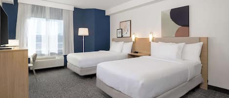 Habitación de hotel con dos camas, un escritorio, una silla, una lámpara y un ventanal con cortinas translúcidas.