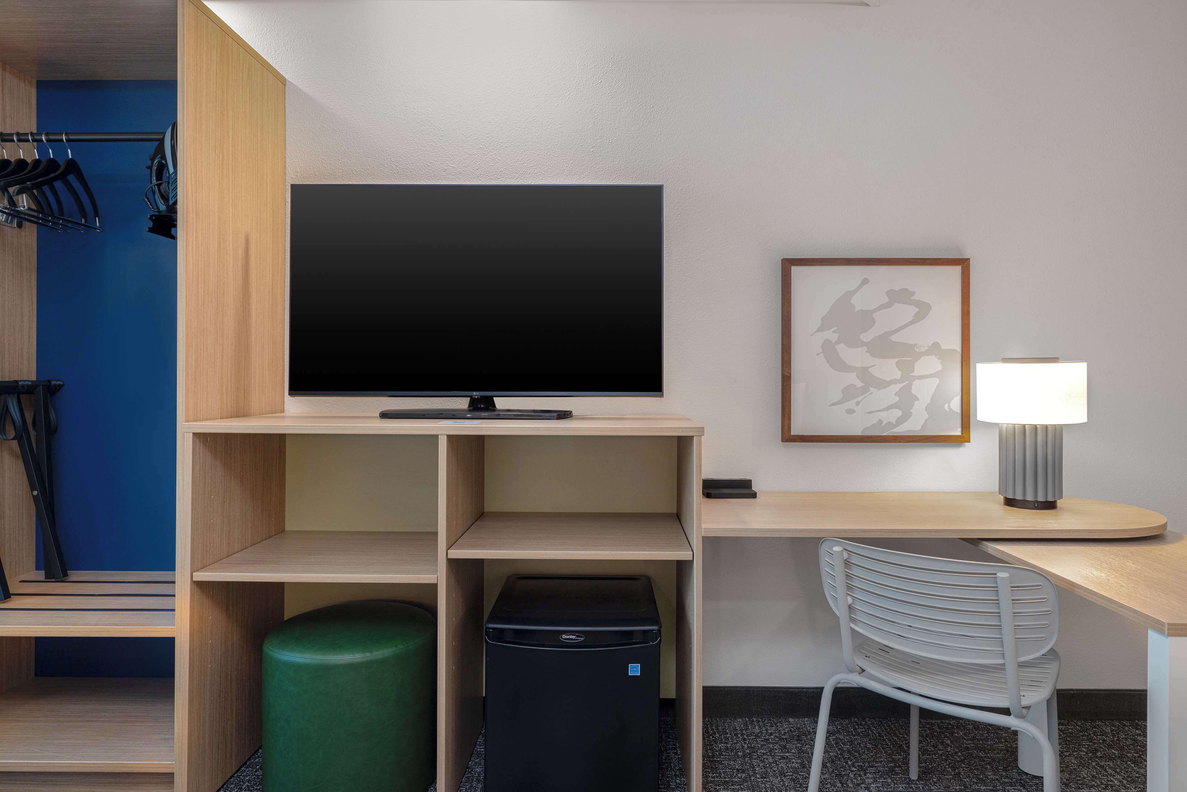 A hotel room with a flat-screen TV, a desk with a lamp, a chair, a trash bin, and a coat rack with clothes.