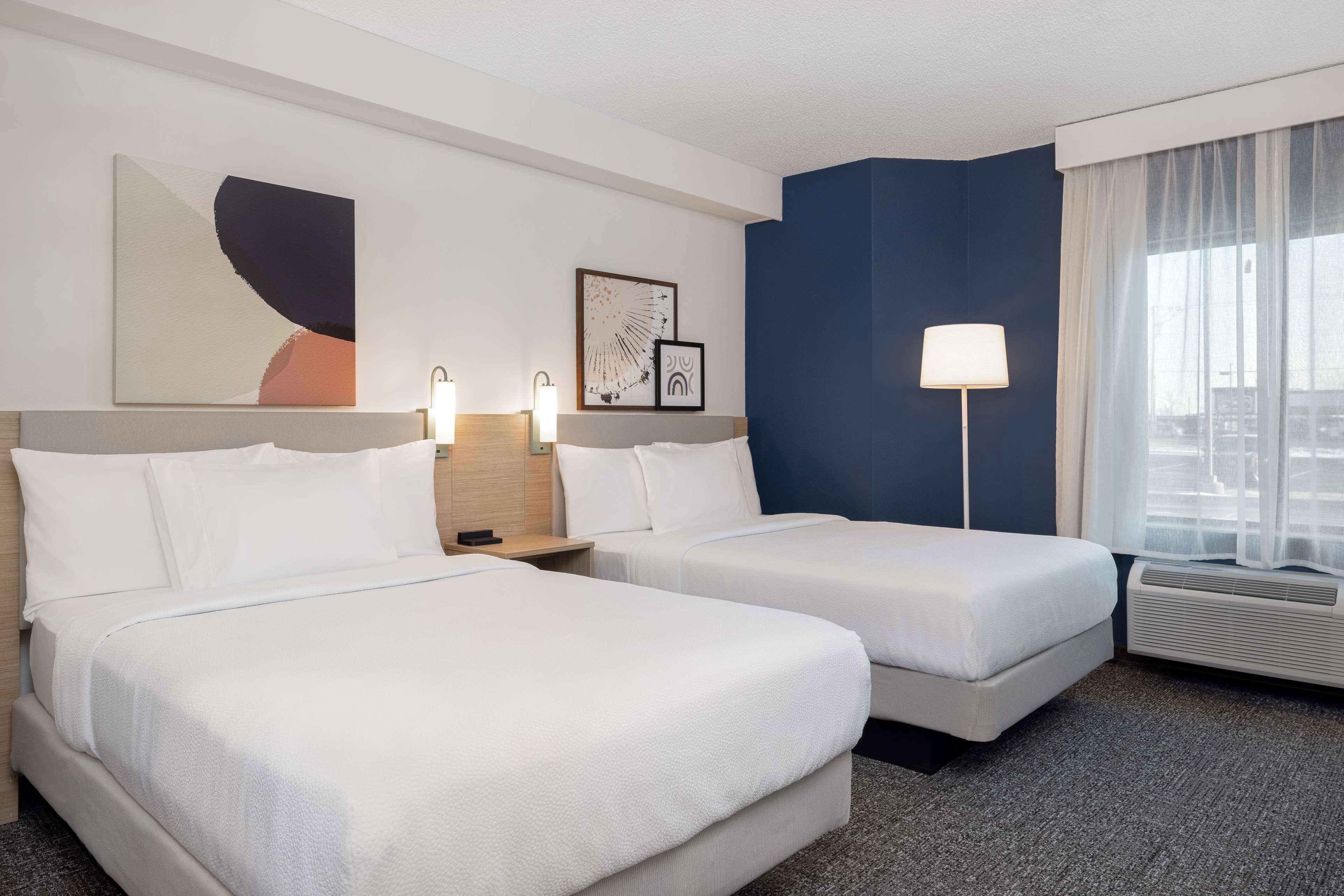 A hotel room with two beds, a large window with sheer curtains, a floor lamp, and artwork on the walls.