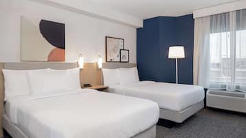 A hotel room with two beds, a large window with sheer curtains, a floor lamp, and artwork on the walls.