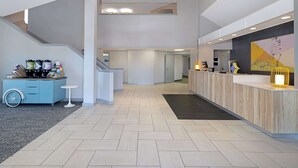 A modern reception area with a light wood reception desk, a small white table, and a blue cart with coffee equipment. - Spark by Hilton Bentonville (Bentonville)