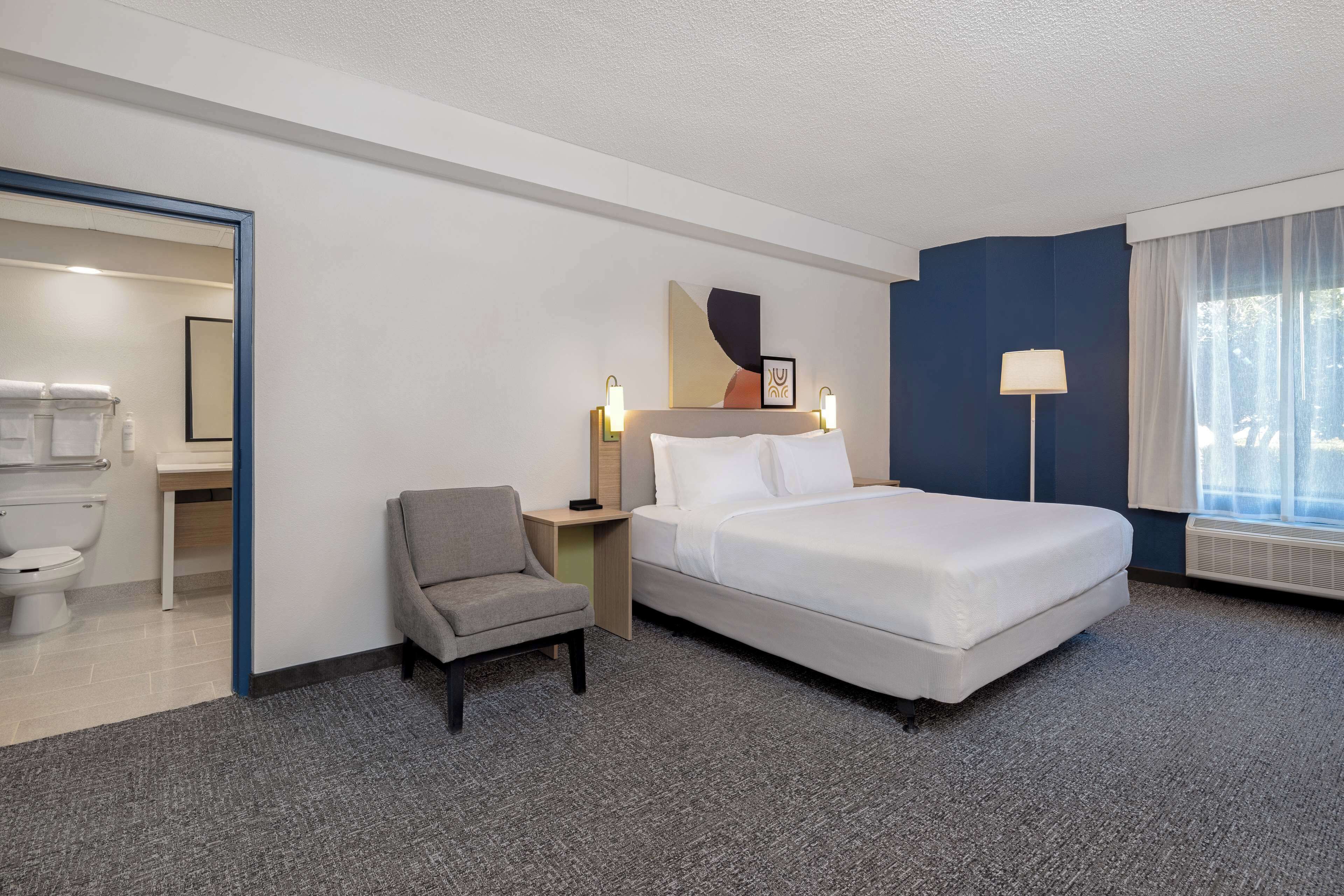Quarto de hotel com cama, uma cadeira, mesa de cabeceira, abajur e janela com cortinas.