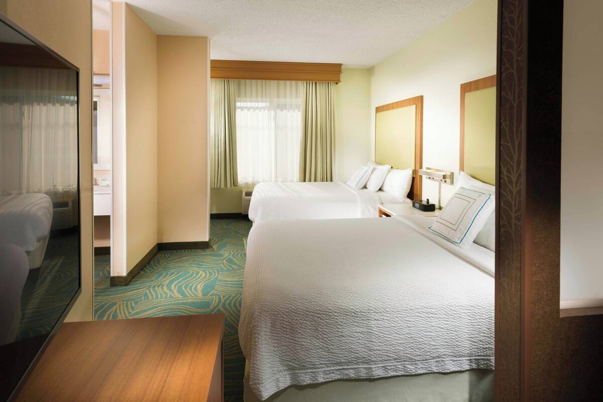 Habitación de hotel con dos camas, una mesita de noche de madera, un espejo grande y una ventana con cortinas.