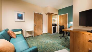 A hotel room with a teal sofa, a small desk, a television, and a kitchenette area.