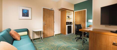 A hotel room with a teal sofa, a small desk, a television, and a kitchenette area.
