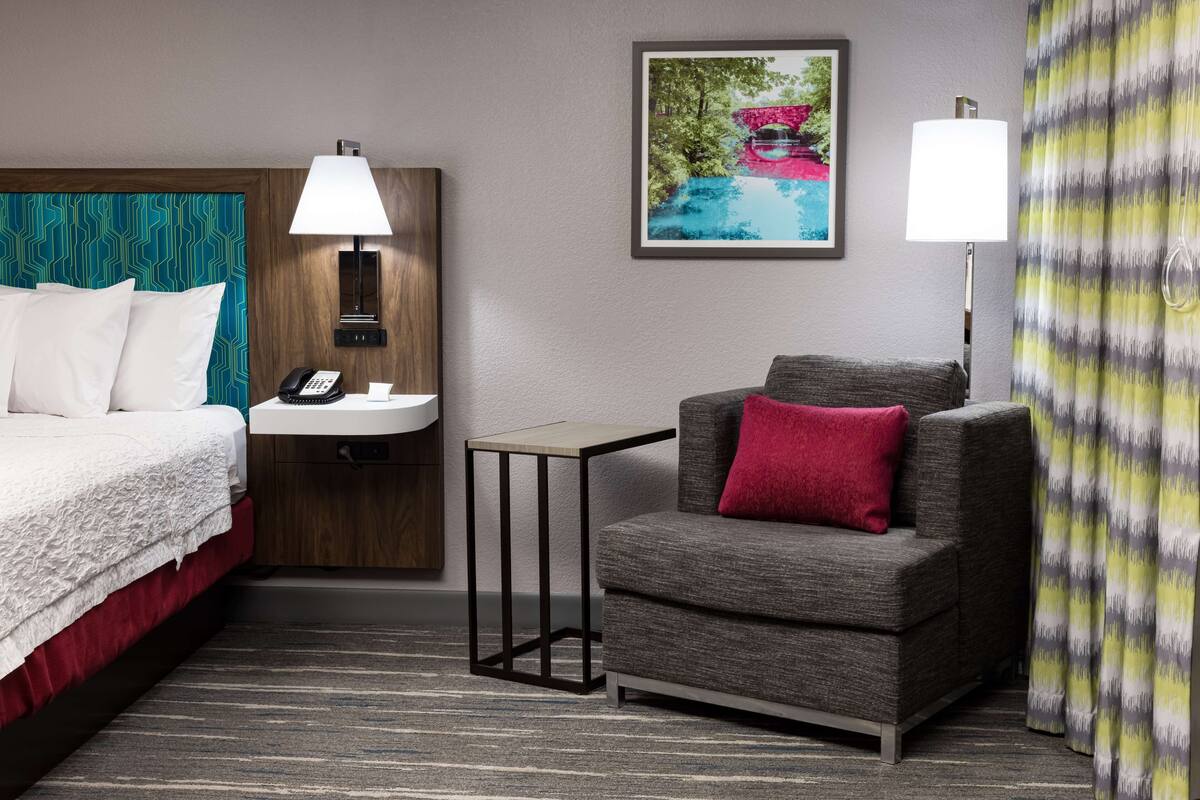 Een hotelkamer met een bed, een klein tafeltje met een lamp, een bank met een rood kussen, een schilderij aan de muur en een kamerkleurig gordijn.
