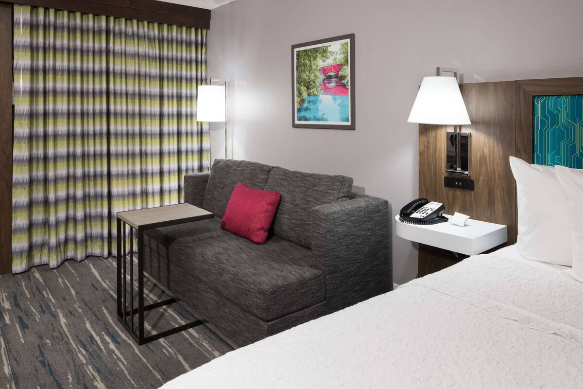 Een hotelkamer met een grijze bank, een tafeltje, een nachtkastje met lamp, een bed en een raam met gordijnen.