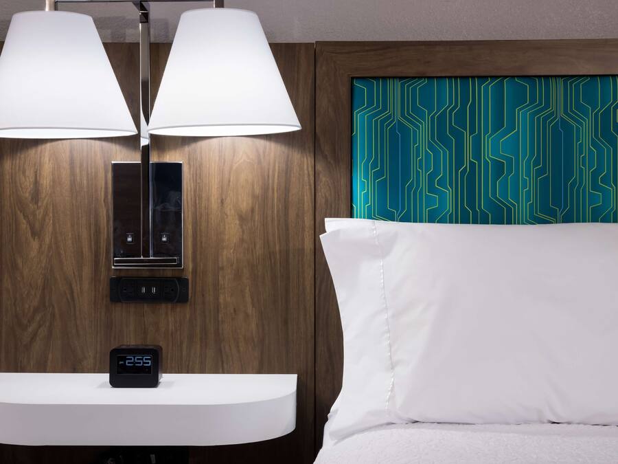 Quarto de hotel com cabeceira de madeira, um relógio digital, mesa de cabeceira e uma luminária.