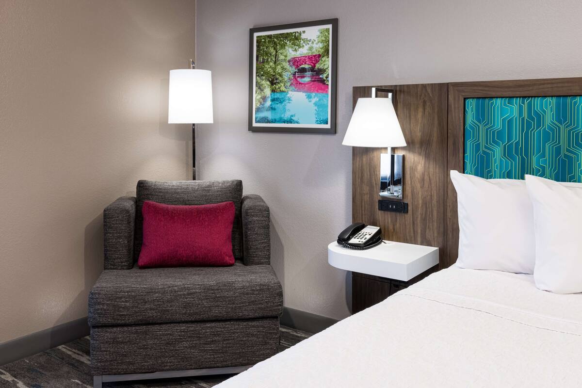 Een hotelkamer met een grijze fauteuil, een wit bed, een nachtkastje met een telefoon en een wandlamp.
