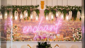 Et værelse med et neonskilt der står "enchant", udsigt over bybilledet, hængende lysarmaturer og en blomsterdekoration.