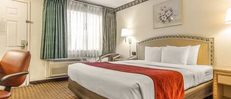 A hotel room with a large bed, a chair, a desk, and a window with curtains.