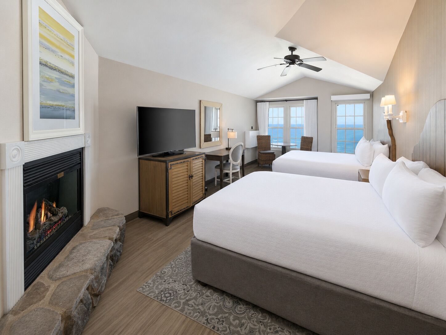 A hotel room with a fireplace, a desk with a television, two beds, a ceiling fan, and a view of the sea.