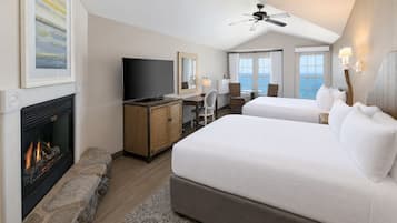 A hotel room with a fireplace, a desk with a television, two beds, a ceiling fan, and a view of the sea.