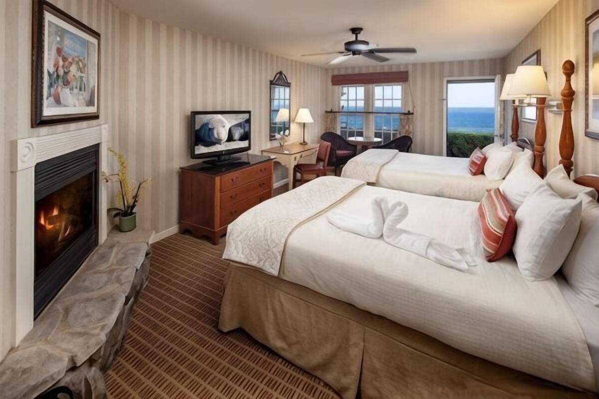 A hotel room with a fireplace, a television, two beds, a desk, and a view of the sea.