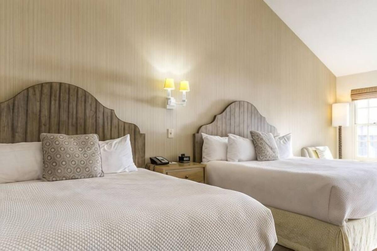 A hotel room with two beds, a nightstand, a lamp, and a wall-mounted light fixture.