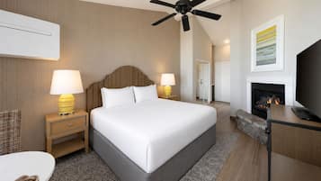 A hotel room with a bed, two bedside lamps, a ceiling fan, a fireplace, a television, and a framed picture on the wall.