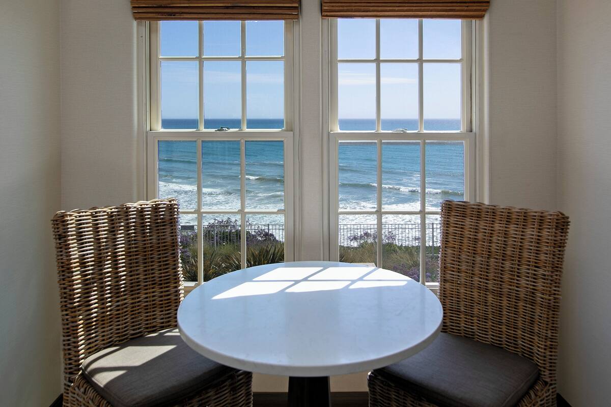A room with a round table and two wicker chairs, a large window with a view of the ocean, and a wooden blind.