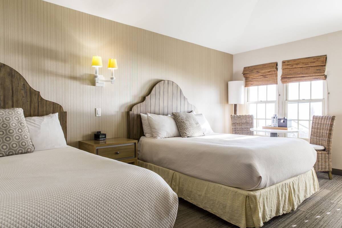 A hotel room with two beds, a nightstand, a lamp, a chair, and a window with blinds.