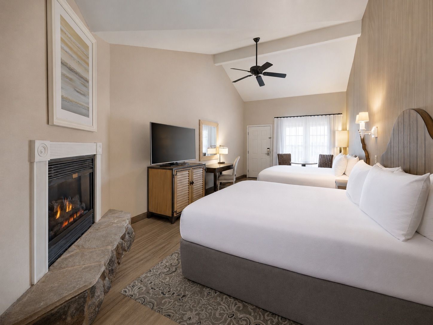 A hotel room with a fireplace, a television, a desk, two beds, and a ceiling fan.
