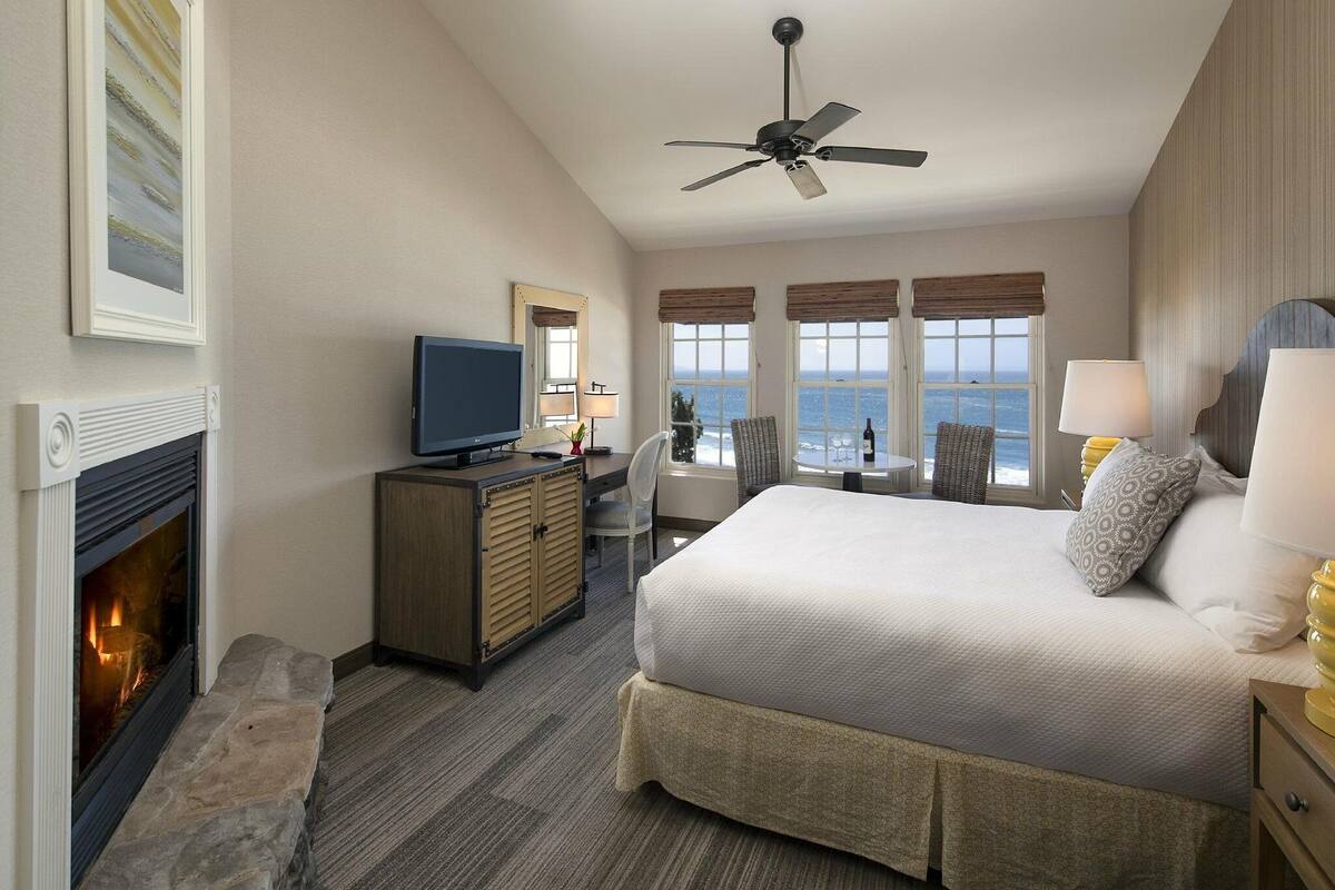 A hotel room with a bed, a television, a fireplace, a desk, and a view of the ocean.