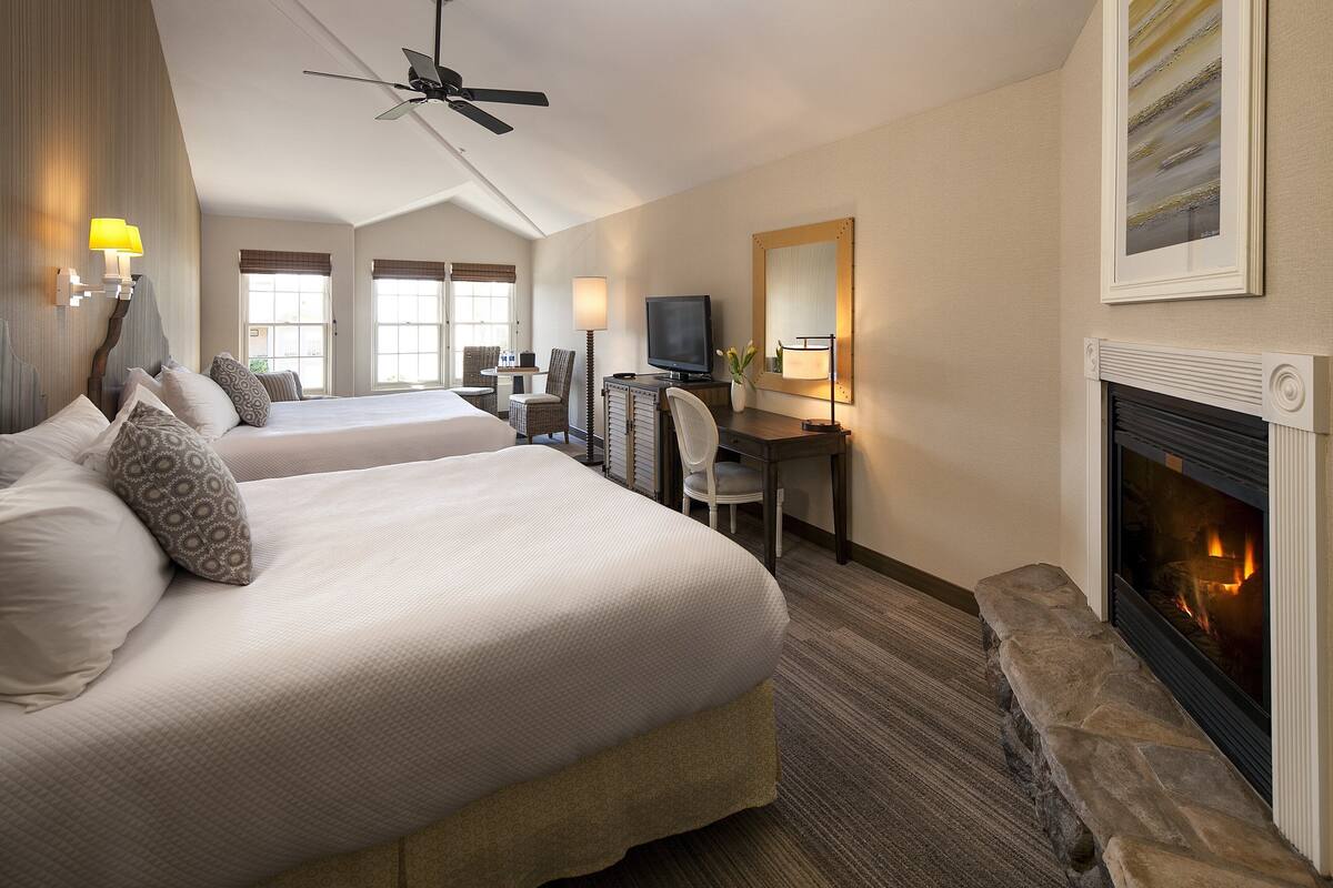 A hotel room with two beds, a desk with a computer, a television, a fireplace, and a ceiling fan.