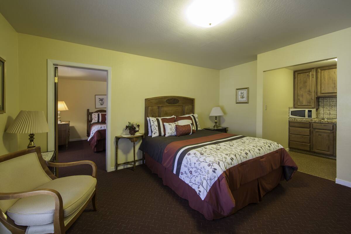 A hotel room with a bed, a chair, a nightstand, a lamp, a picture frame, and a kitchen area.