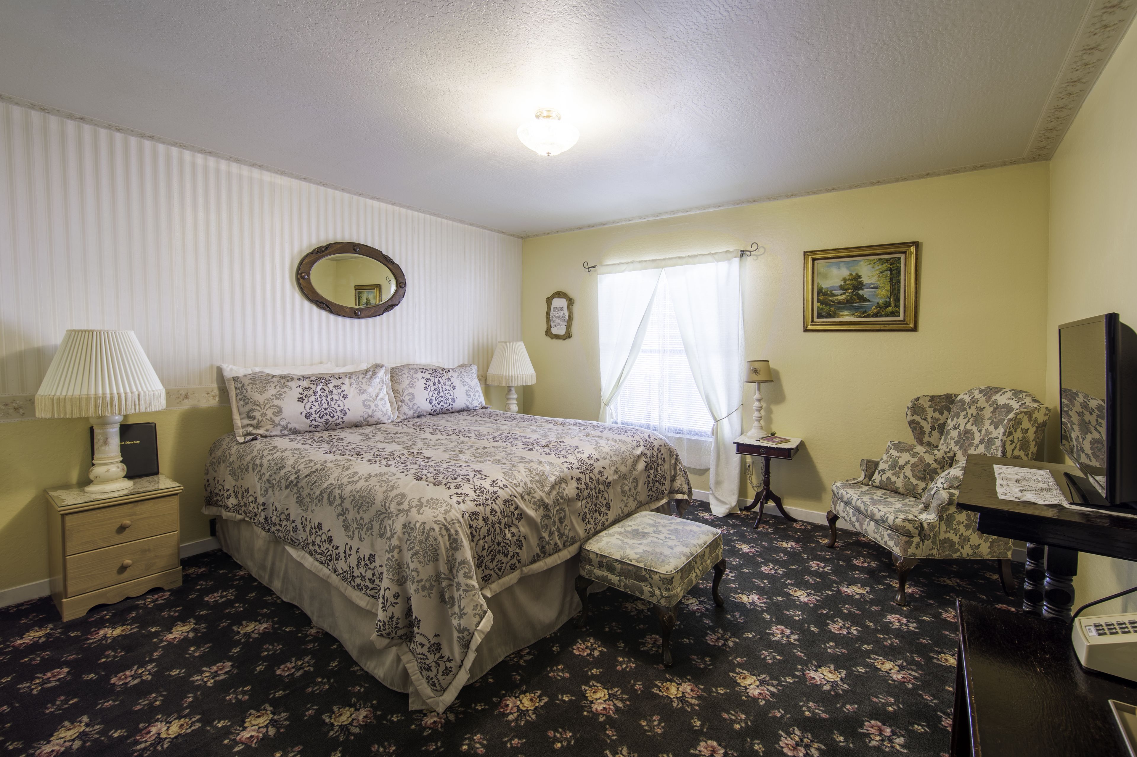 A bedroom with a bed, bedside tables, a chair, a desk, a lamp, a mirror, a window with curtains, and a painting on the wall.