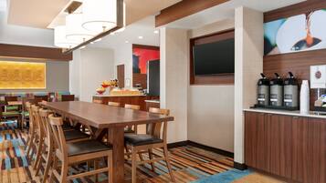 Un espace repas d’hôtel avec une longue table en bois, de nombreuses chaises, un coin café et un téléviseur fixé au mur.