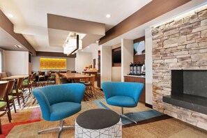 A modern hotel lobby with a stone fireplace, blue armchairs, a dining area with wooden tables and chairs, and a large TV mounted on the wall. - Fairfield Inn by Marriott Roseville (Roseville)