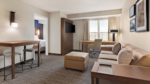 A hotel room with a desk, two chairs, a sofa, an ottoman, a flat-screen TV, and framed pictures on the wall.
