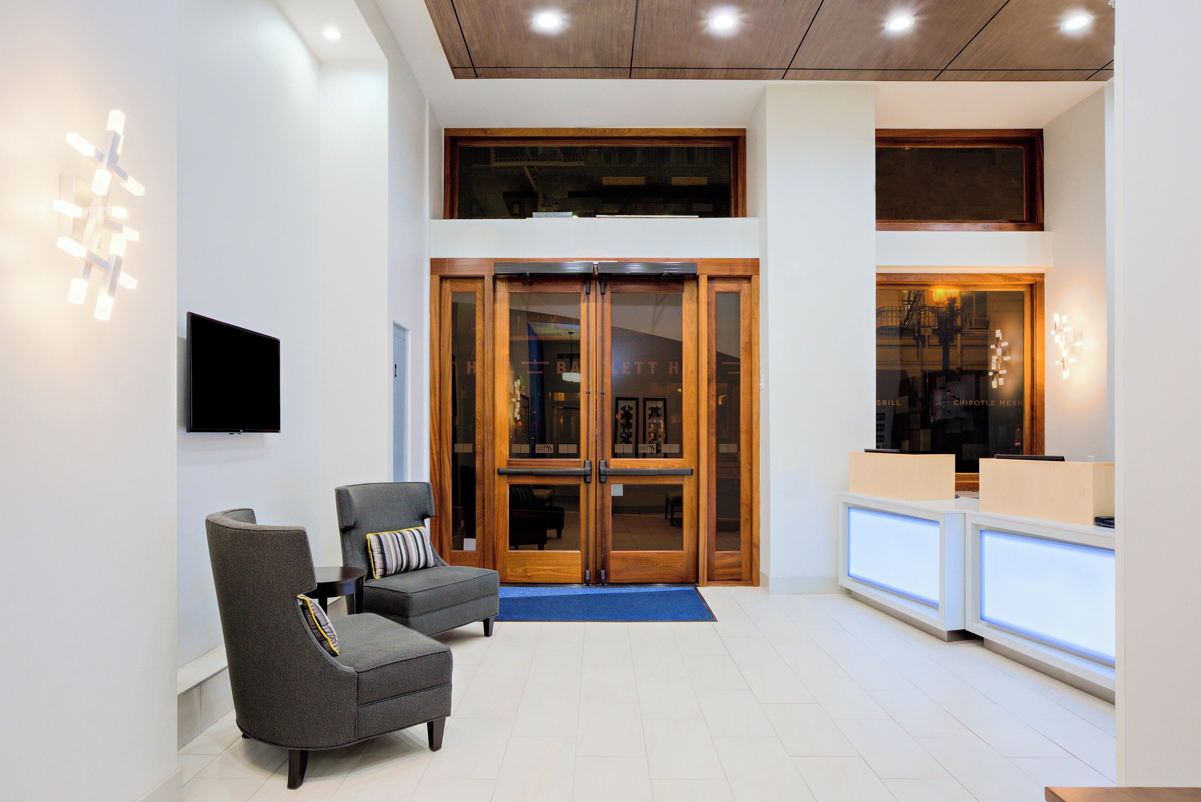 A lobby with a reception desk, two armchairs, a flat-screen TV, and a wooden door leading to a restaurant.