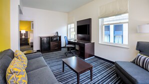 A hotel room with a gray sofa, a small table, a desk with a chair, a TV, and a window with blinds. - Holiday Inn Express San Francisco Union Square by IHG (San Francisco)