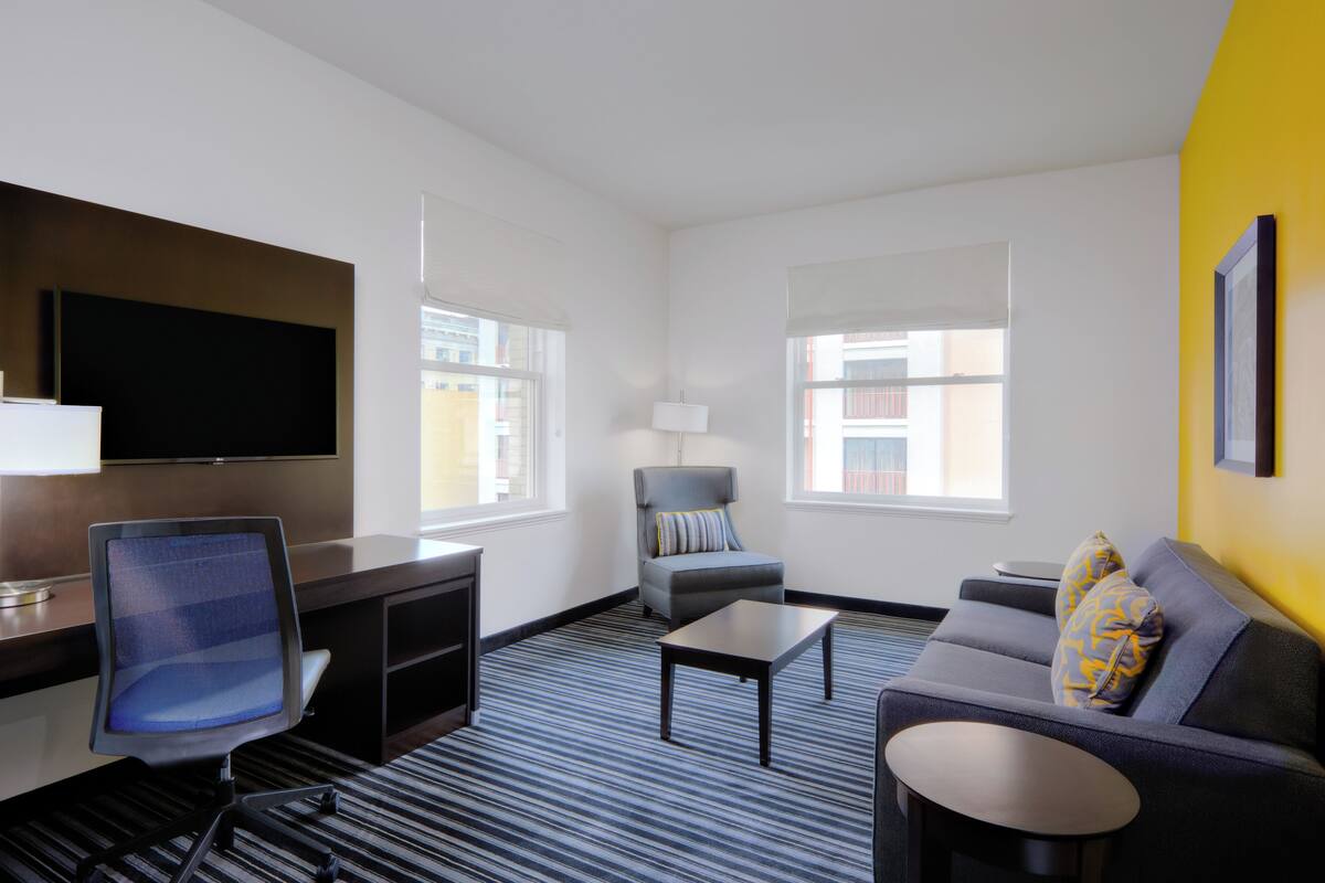 A hotel room with a flat-screen TV, a desk, a chair, a sofa, and two windows.