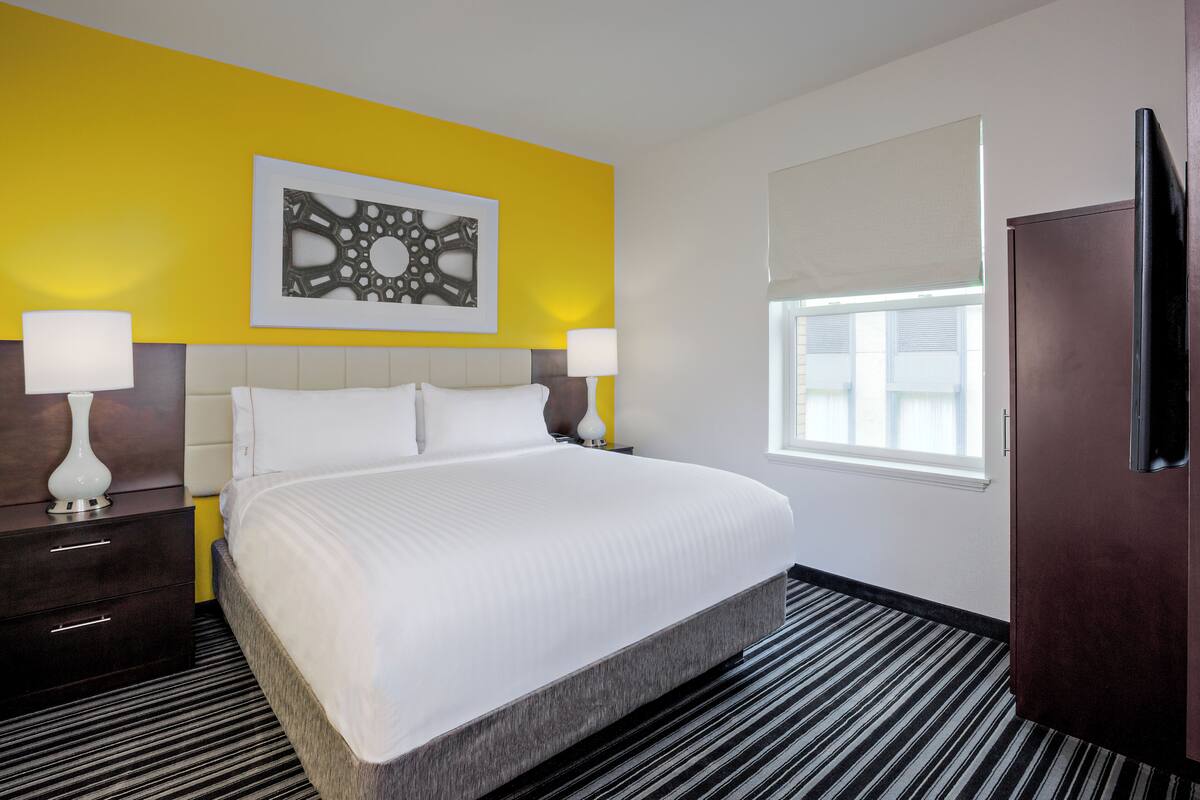 A hotel room with a large bed, two bedside tables with lamps, a window with blinds, and a wall-mounted TV.