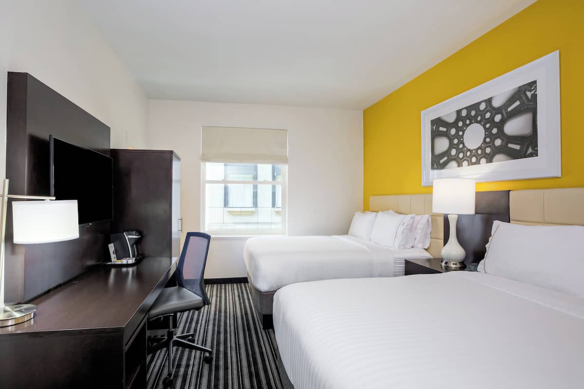 A hotel room with two beds, a desk with a chair, a TV, and a window with blinds.