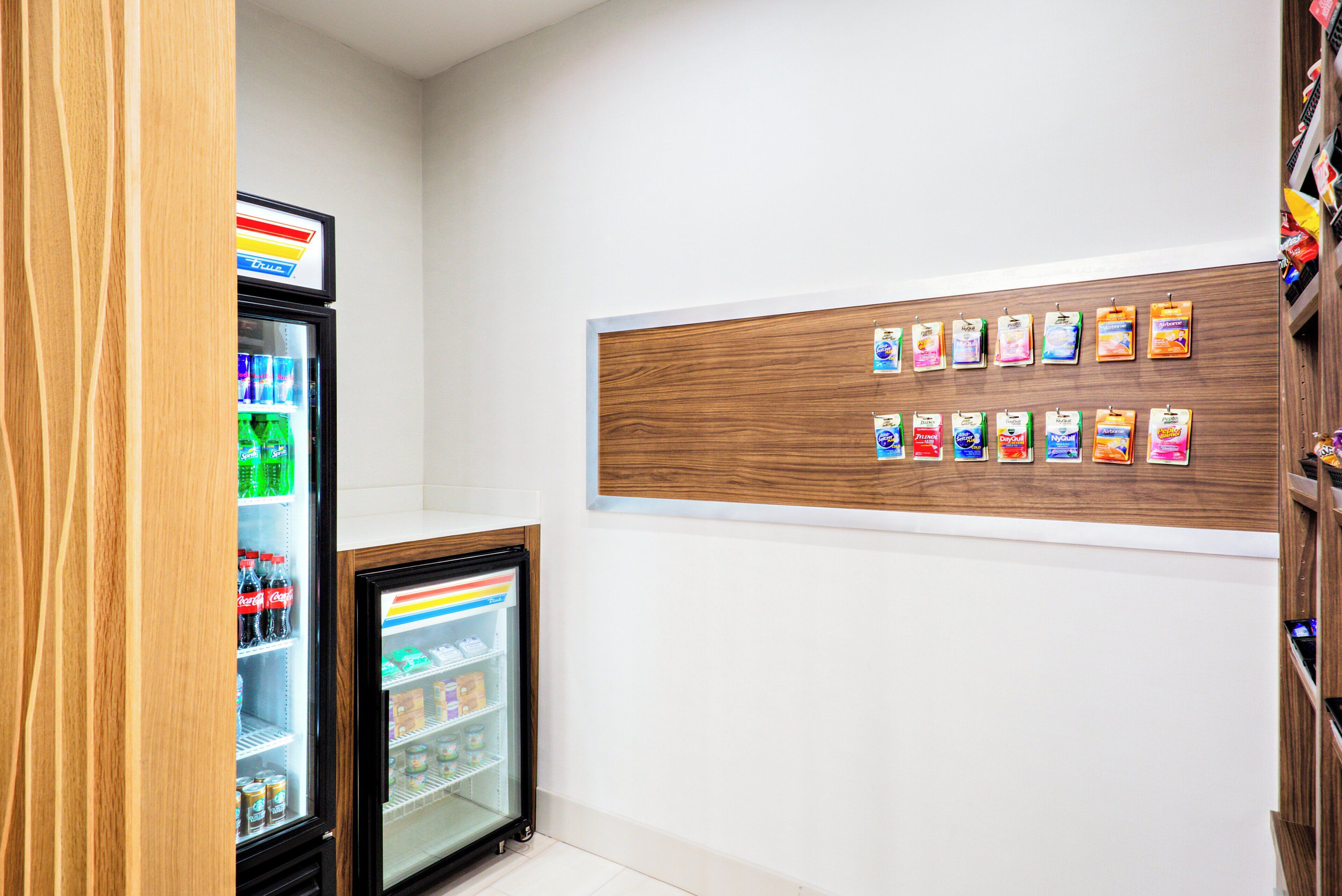 Uma pequena área comercial com vitrine refrigerada e painel de madeira com embalagens de produtos penduradas.
