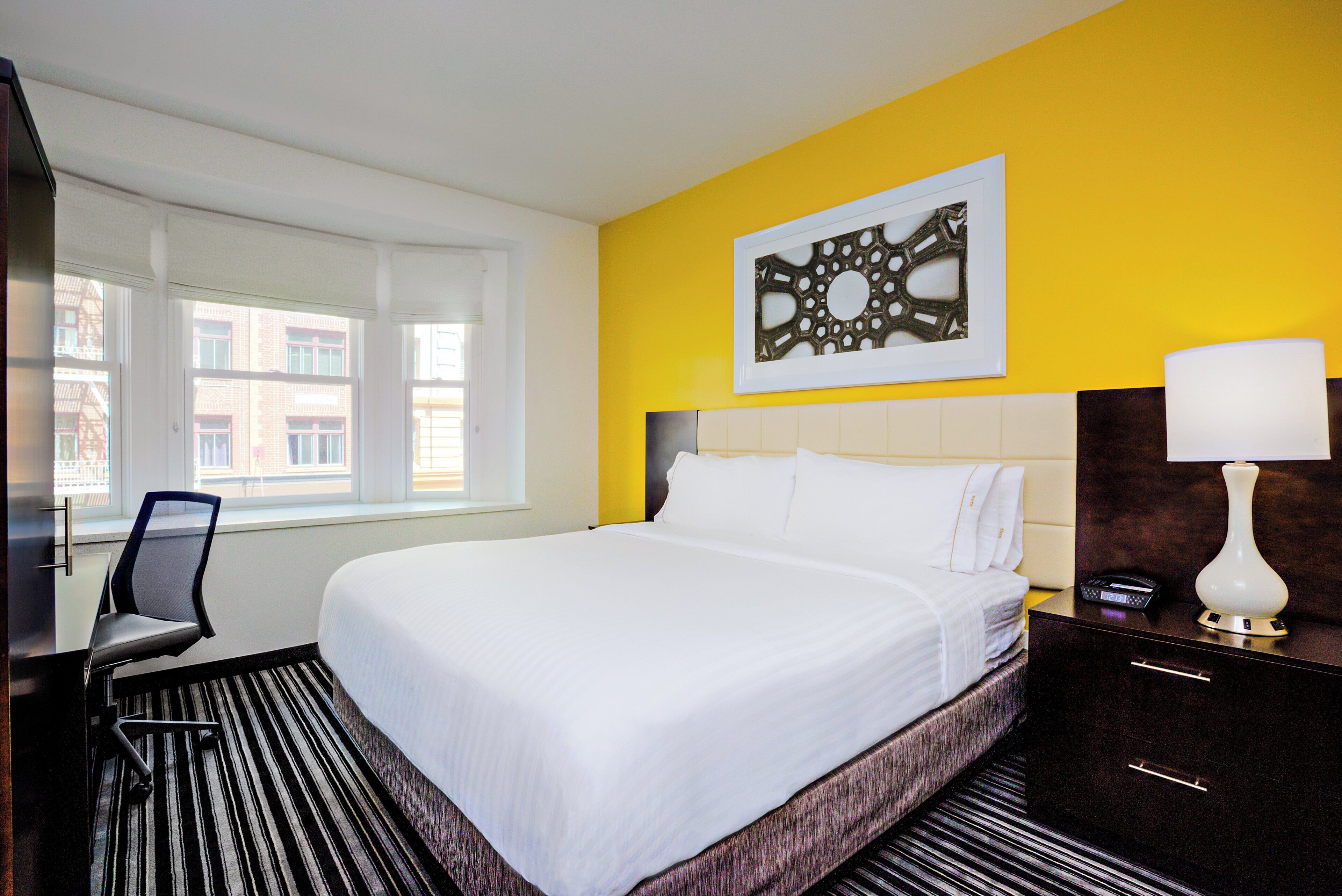 A hotel room with a bed, a chair, a desk, a lamp, and a window with a view of buildings.